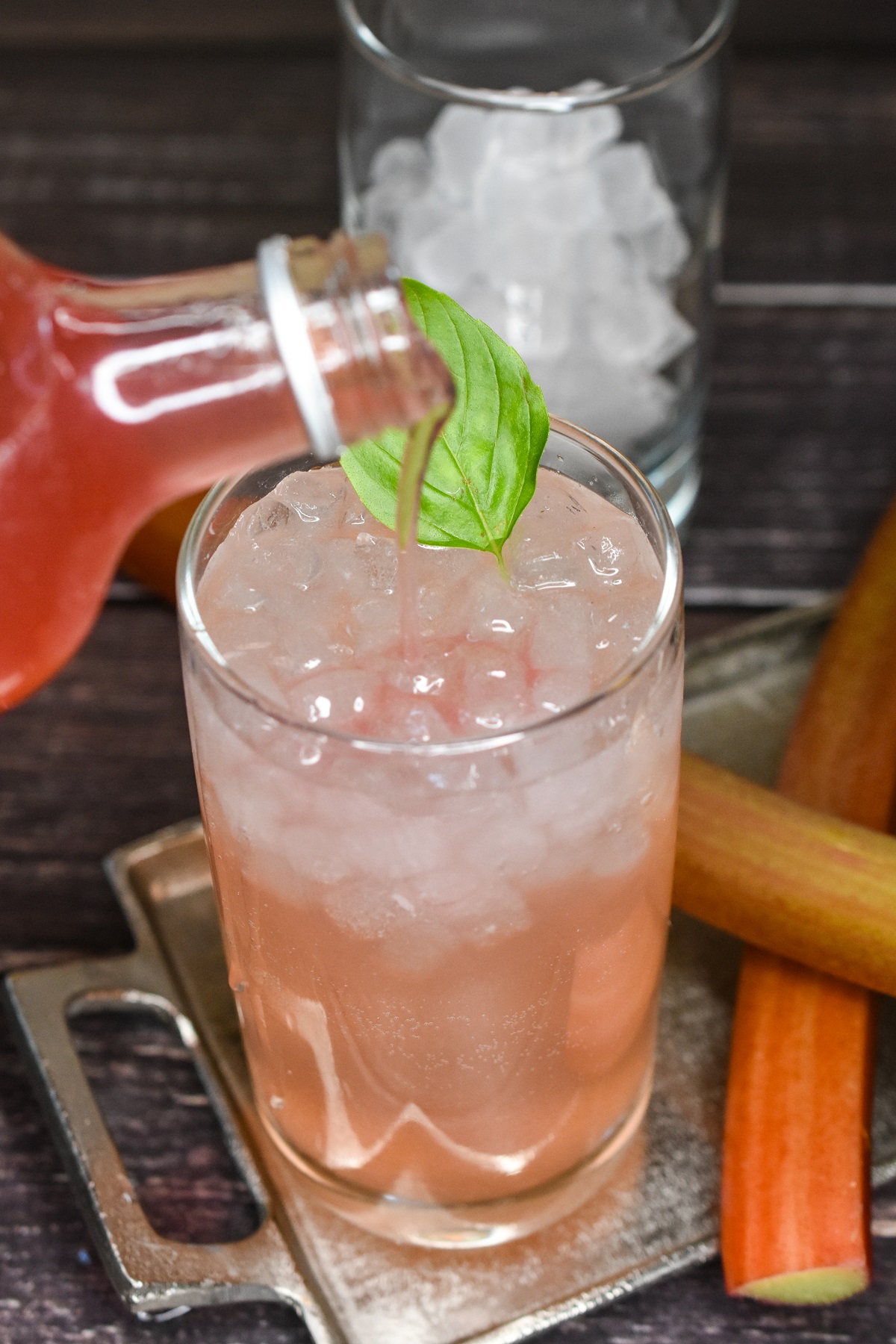 Easy Homemade Rhubarb Syrup recipe. Shown in a glass bottle being poured into a glass full of ice and club soda to make Rhubarb Soda. Garnished with basil leaf