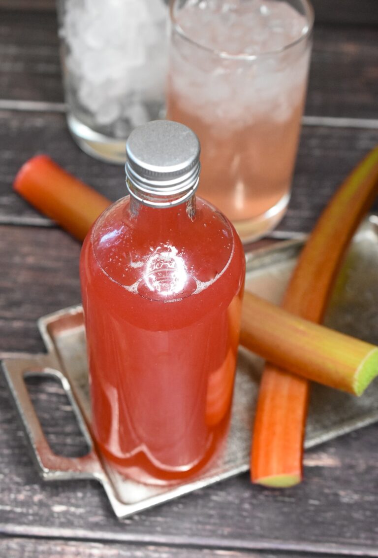 Homemade Rhubarb Syrup in a glass bottle alongside two stalks of fresh rhubarb with Rhubarb soda in background