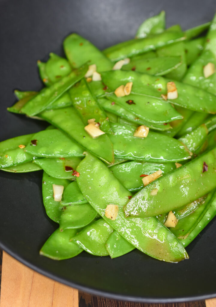 Sautéed Snow Peas shown on a black plate. Speckled with chopped garlic and red pepper flakes