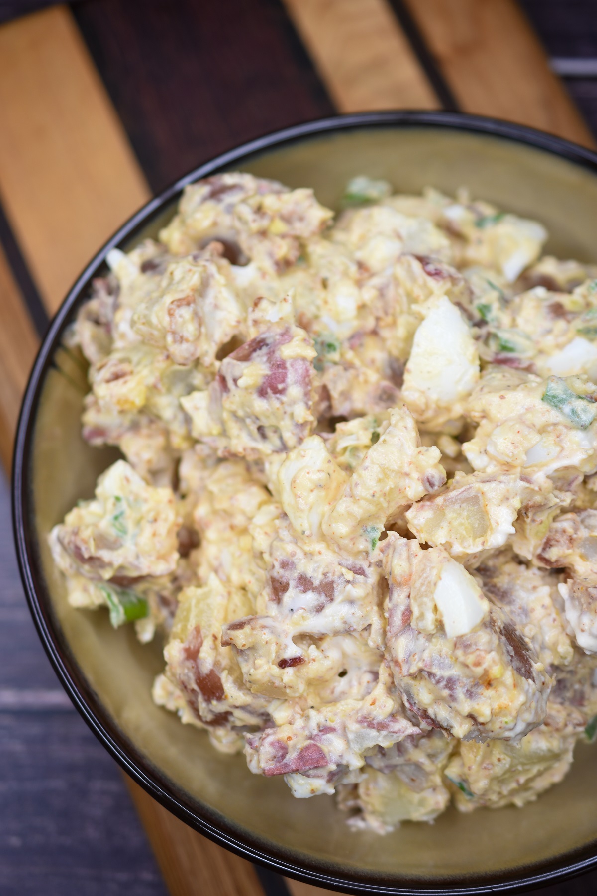 potato salad made with red potatoes, mayo, mustard, scallions and bacon shown in a bowl with brown rim on a wood cutting board