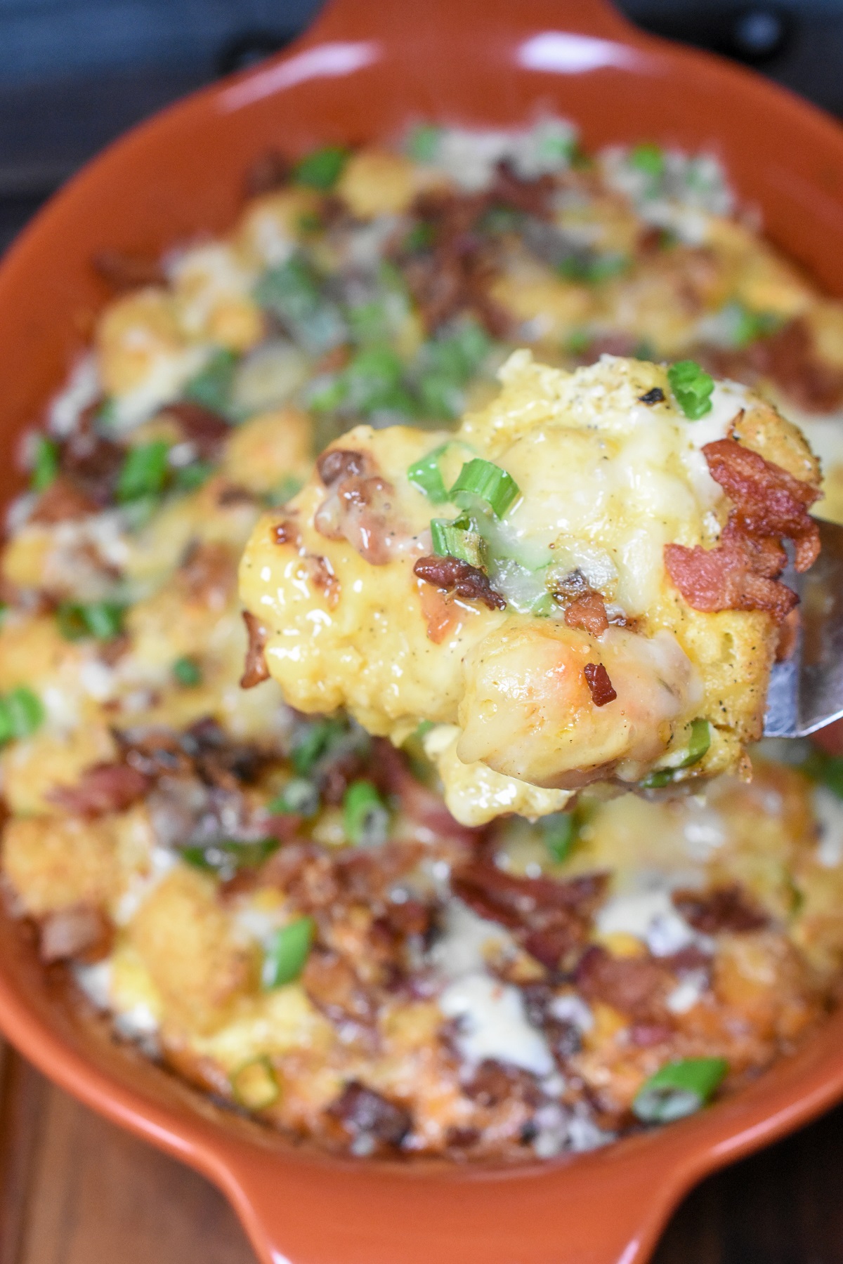 Tater Tot Breakfast Casserole. Shown in an orange casserole dish with some held above it on a spoon. Garnished with scallions, bacon and cheese