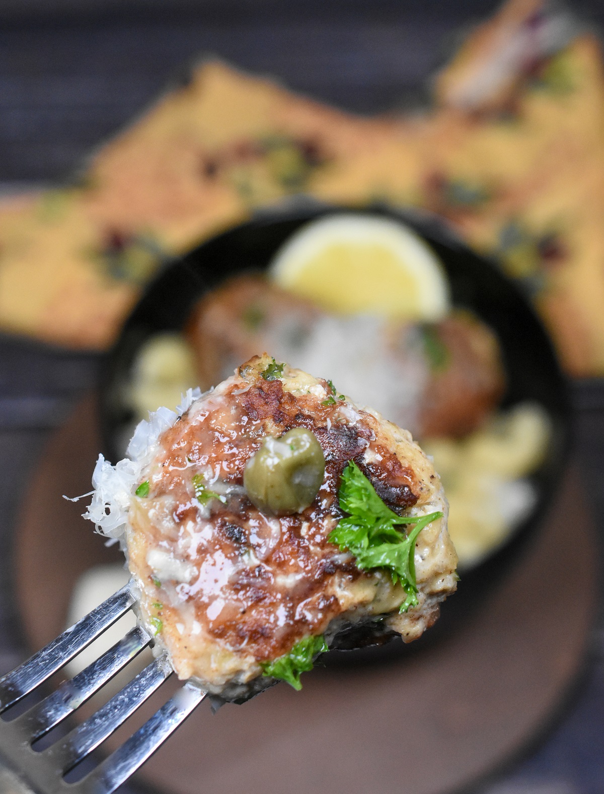 A Chicken Piccata Meatball on a fork with a little fresh grated Parmesan cheese, parsley and a caper, glazed in Piccata Sauce.