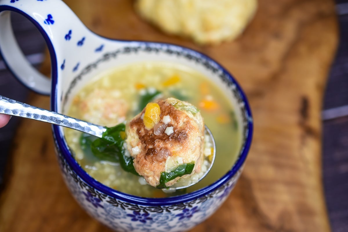 chicken pastina soup with mini chicken meatballs shown in a Polish pottery soup bowl with curved handle that is white with blue accents. A spoon hovering over holds some of the soup and a mini chicken meatball.